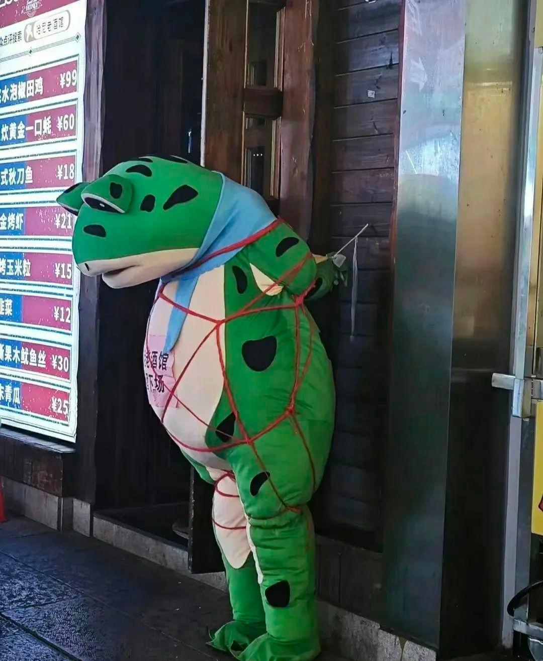 一个人穿着青蛙玩偶服被绑在门上,让人不解.是被人欺负了?