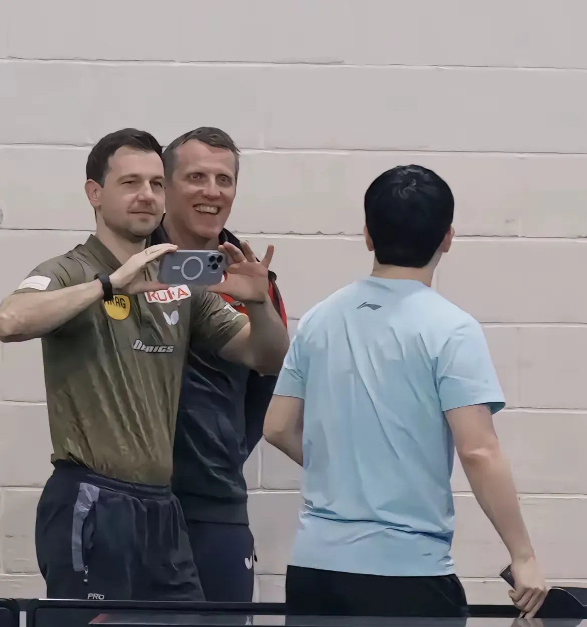 波尔开心的拿着手机给好友樊振东拍照,德国队教练罗斯科夫也高兴的合