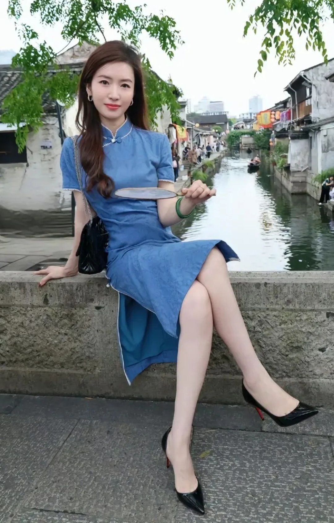 来一组江南水乡穿搭,蓝色旗袍搭配白色开衫,手拿小团扇,在小桥流水的