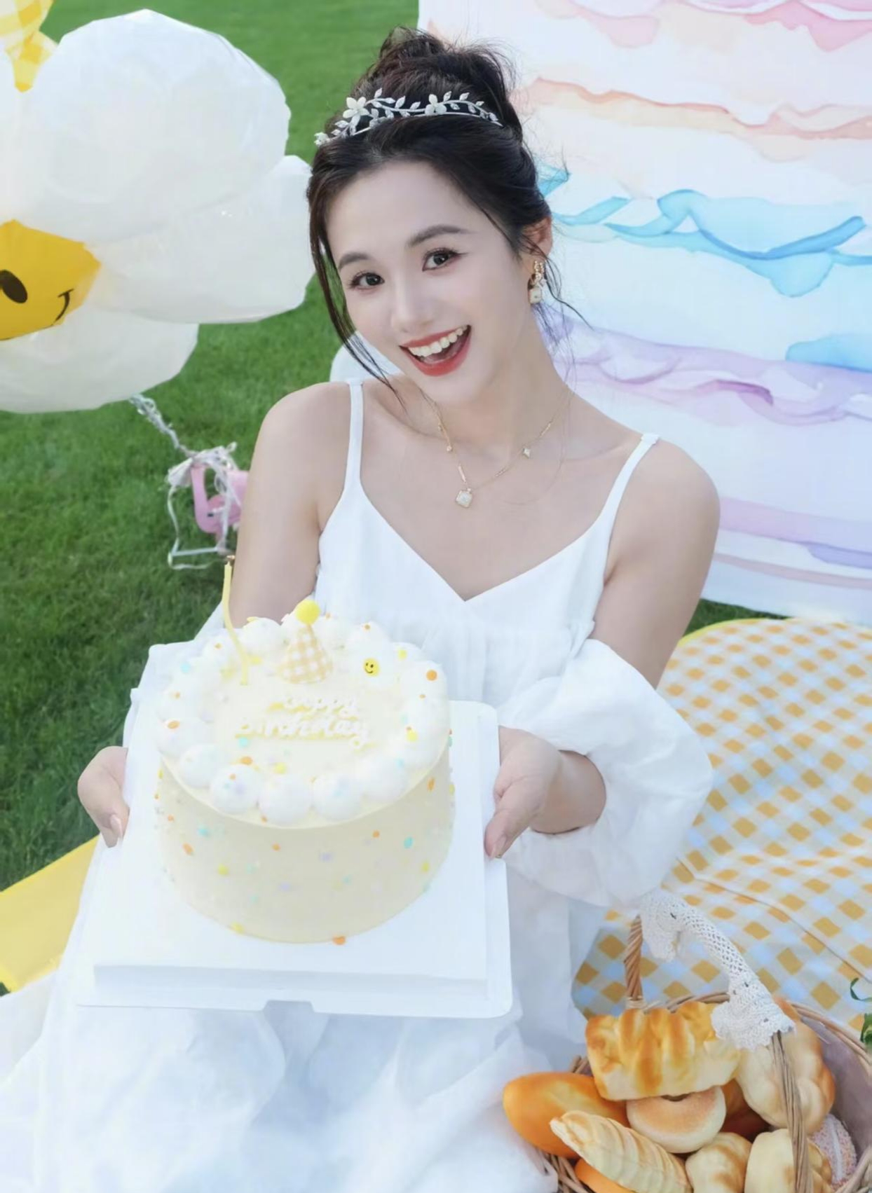 安悦溪身穿白色长裙,手持精致蛋糕,笑容灿烂,在户外的草坪上,阳光洒下