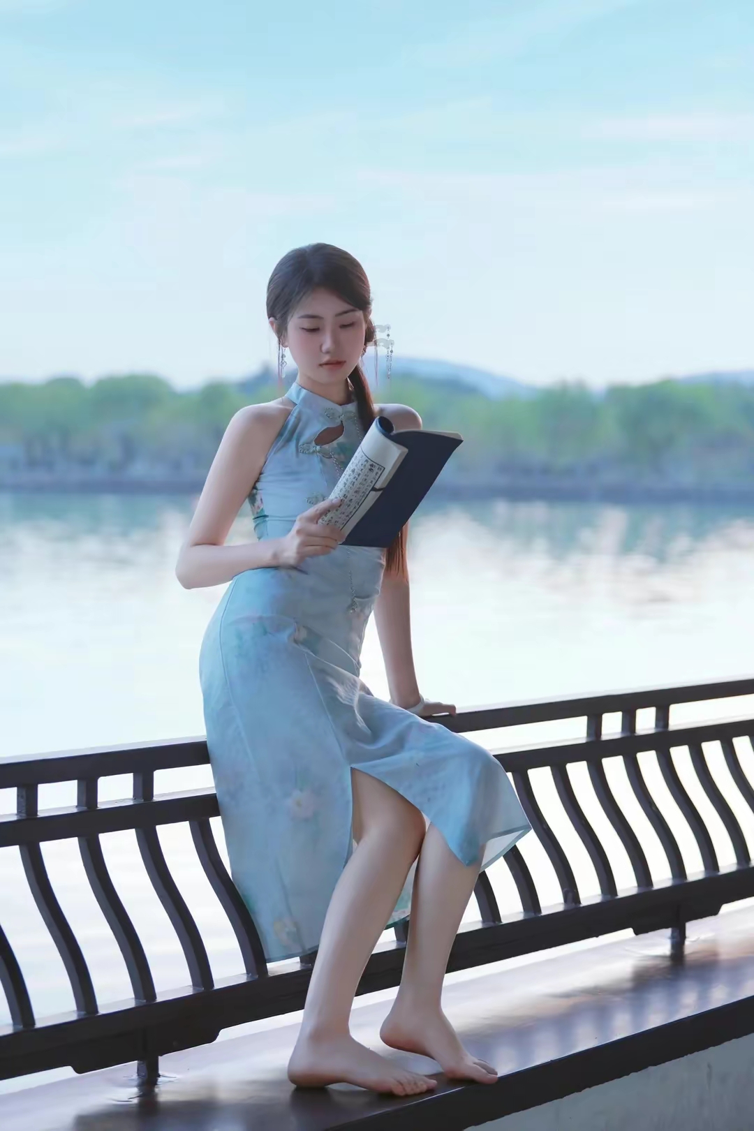 一个穿着旗袍的美女,坐在湖边的小亭子里,一边欣赏美景,一边翻看书籍