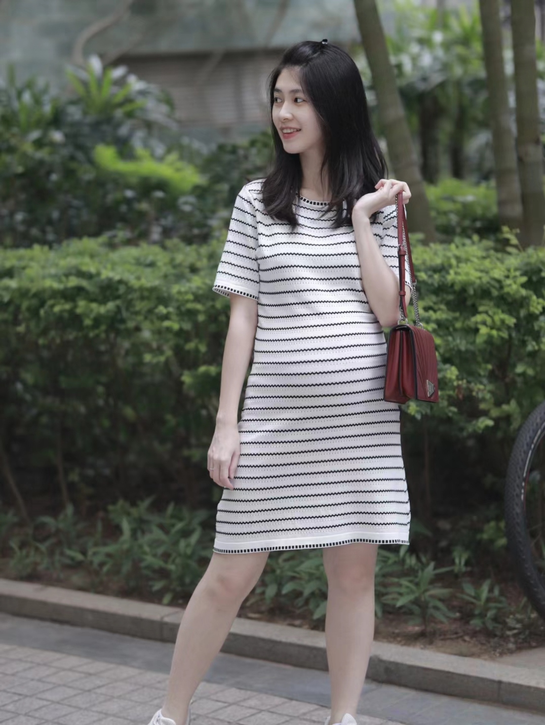 穿着条纹裙的小姐姐,笑容甜美,手提小红包,漫步在公园里,享受着悠闲的