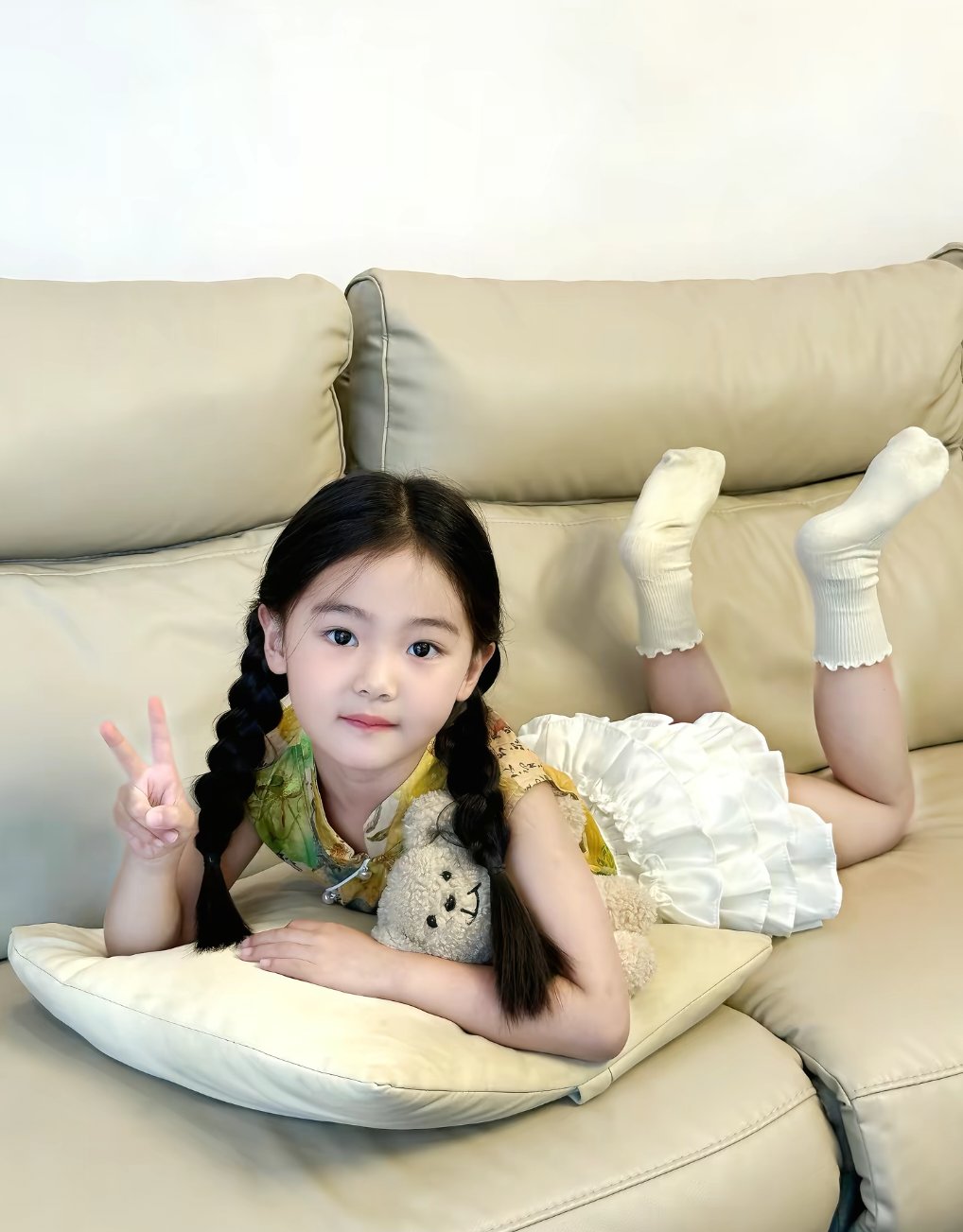 小女孩躺在沙发上,手比着和平标志,旁边是她的泰迪熊