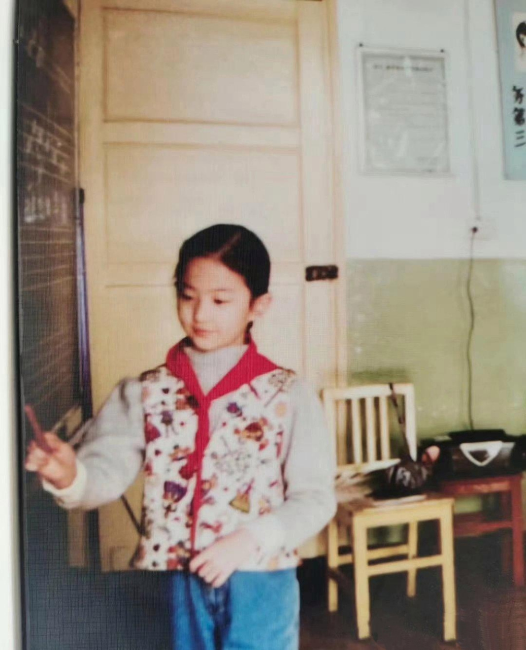 刘亦菲童年的一张旧照,她身穿花夹克,胸前戴着红领巾,站在黑板前手指